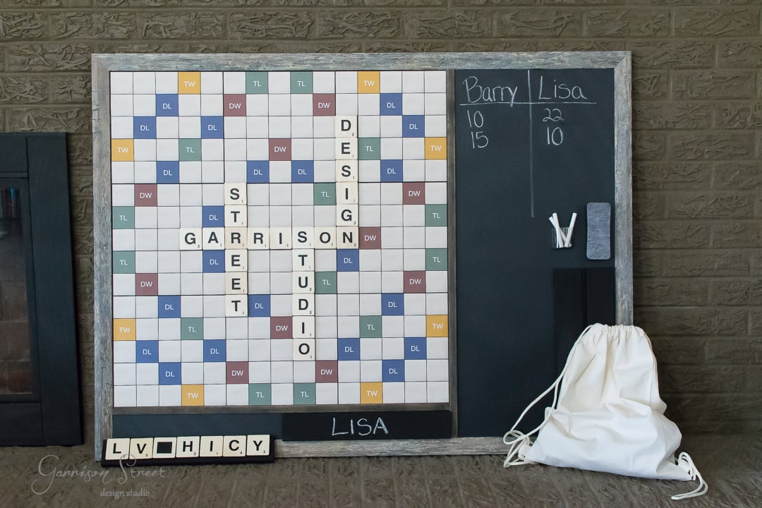 A large Scrabble board with tiles spelling "Garrison Street Design Studio" is framed by a chalkboard. Names "Barry" and "Lisa" with scores are written on the board. Nearby, there's a small bag and letter rack with tiles spelling "L V H I C Y.