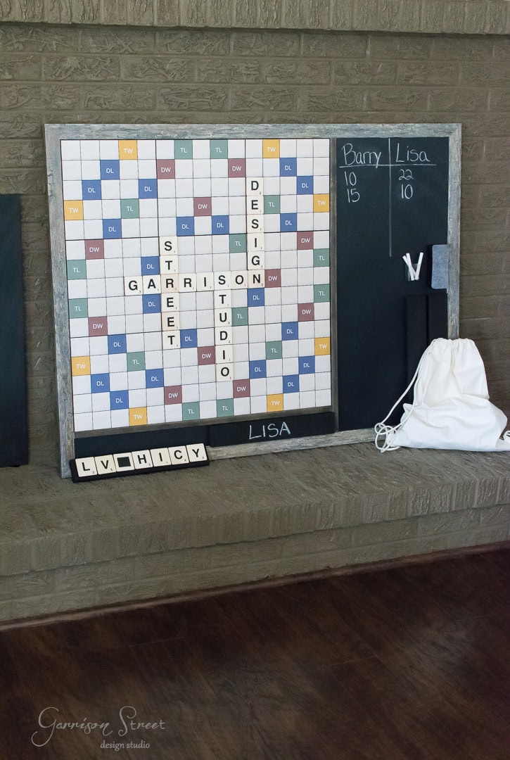 A large Scrabble board is mounted on a wall next to a chalkboard with scores for Barry and Lisa. Scrabble tiles spell out "Garrison Street Design Studio." A white bag and tiles spelling "LVHICY" are also visible.