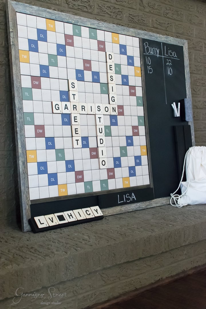 A large Scrabble board is framed on a brick wall, displaying words like "Garrison," "Street," "Design," and "Studio." Below, a scoreboard lists names and scores. Scrabble letters "L," "V," "H," "C," "Y" are on a holder. A white bag sits nearby.