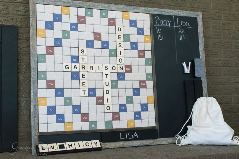 A large, Scrabble-themed game board displays words like "GARRETT," "STUDIO," "DESIGN," and "STREET." A chalkboard beside it shows scores for Barry and Lisa. A white bag and a rack with tiles are on the right.
