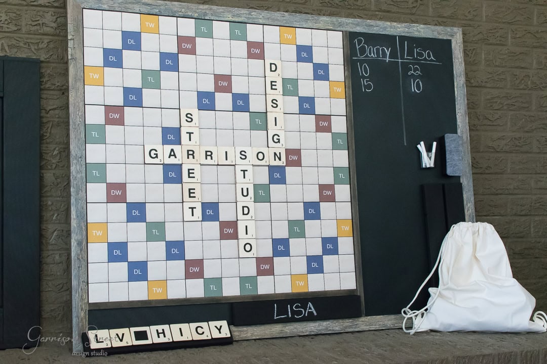 A large, Scrabble-themed game board displays words like "GARRETT," "STUDIO," "DESIGN," and "STREET." A chalkboard beside it shows scores for Barry and Lisa. A white bag and a rack with tiles are on the right.