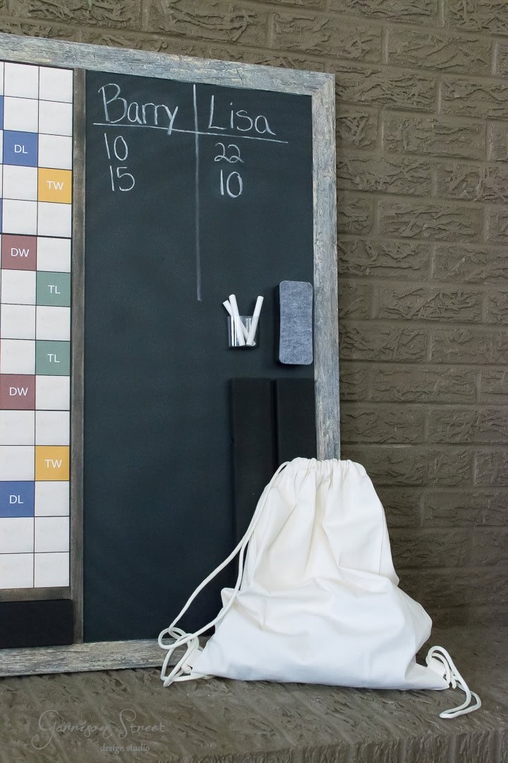 A chalkboard with scores listed for Barry and Lisa. The score for Barry is 10 and 15, while Lisa's score is 22 and 10. Chalk and an eraser rest on a tray, and a white drawstring bag is placed nearby against a brick wall.