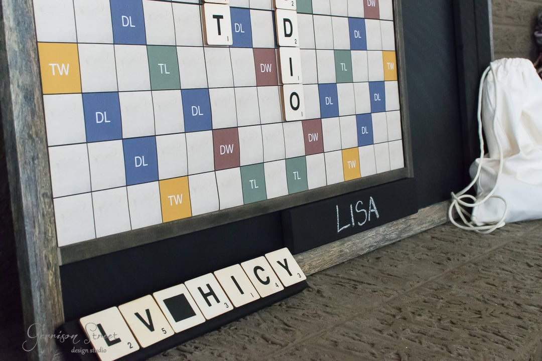 A Scrabble board with tiles spelling "TUDIO". Nearby, a tile rack displays the letters "V H I C Y". The name "Lisa" is written on the board's base, and a white drawstring bag is placed beside it.