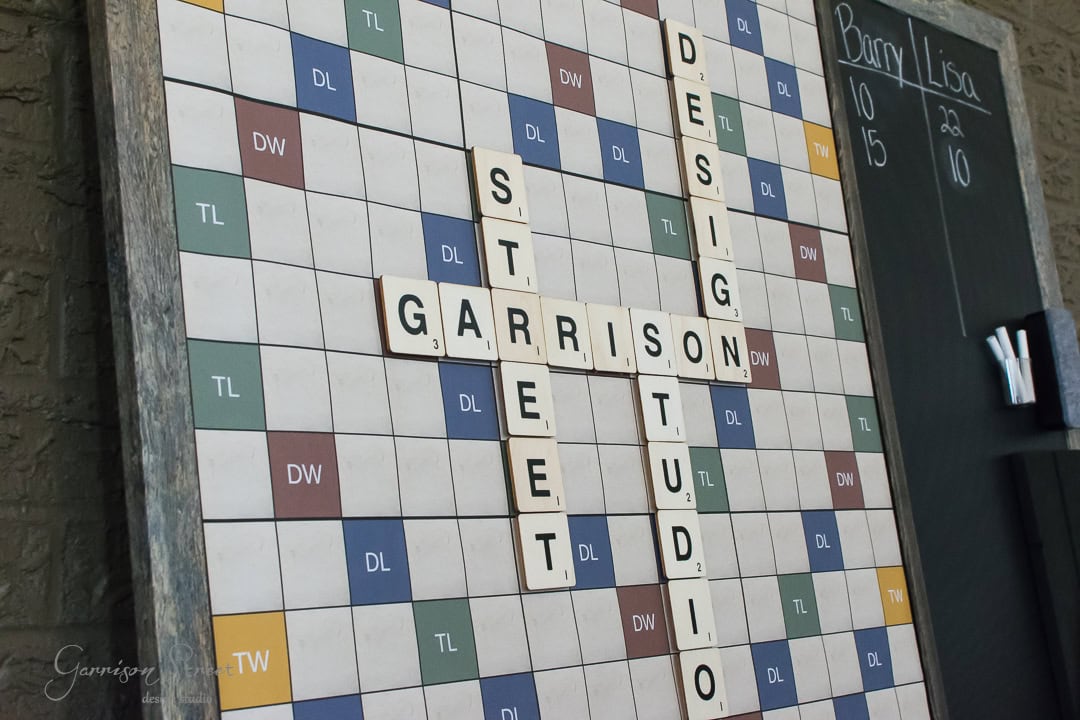 A large Scrabble board with the words "Garrison Street Design Studio" arranged in the center. To the right is a chalkboard with headings "Barry" and "Lisa" and scores 10, 15, and 20. A small rack holds the letter "W".