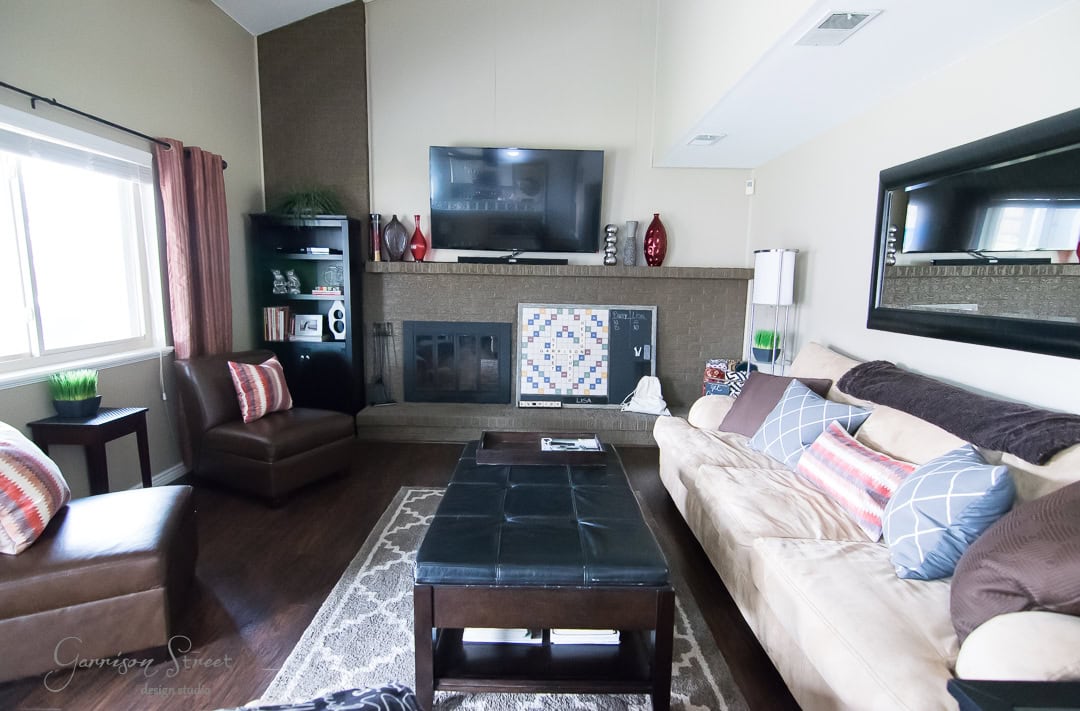 A modern living room with a large beige sofa, several patterned pillows, and a matching armchair. A flat-screen TV is mounted above a fireplace. Shelves with decor items flank the fireplace. A coffee table sits on a geometric rug on wooden flooring.