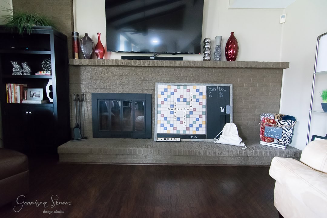 A living room features a large brick fireplace with a mounted TV above. Decor includes vases, books, and a scrabble board. A dark wooden floor contrasts with the light walls, and a black bookshelf displays various items on the left.
