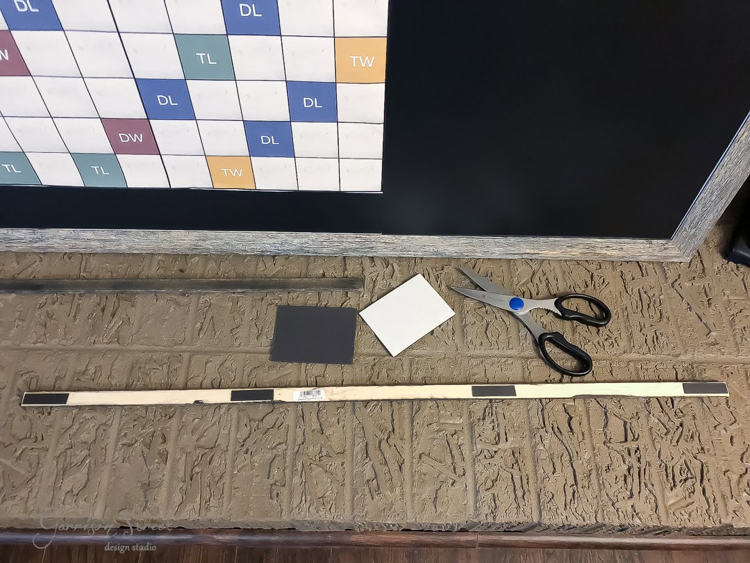 A table with crafting materials, including a long wooden strip with magnets, a small black square, a white square, black scissors, and a chart on a black board in the background with various letters in different colored squares.