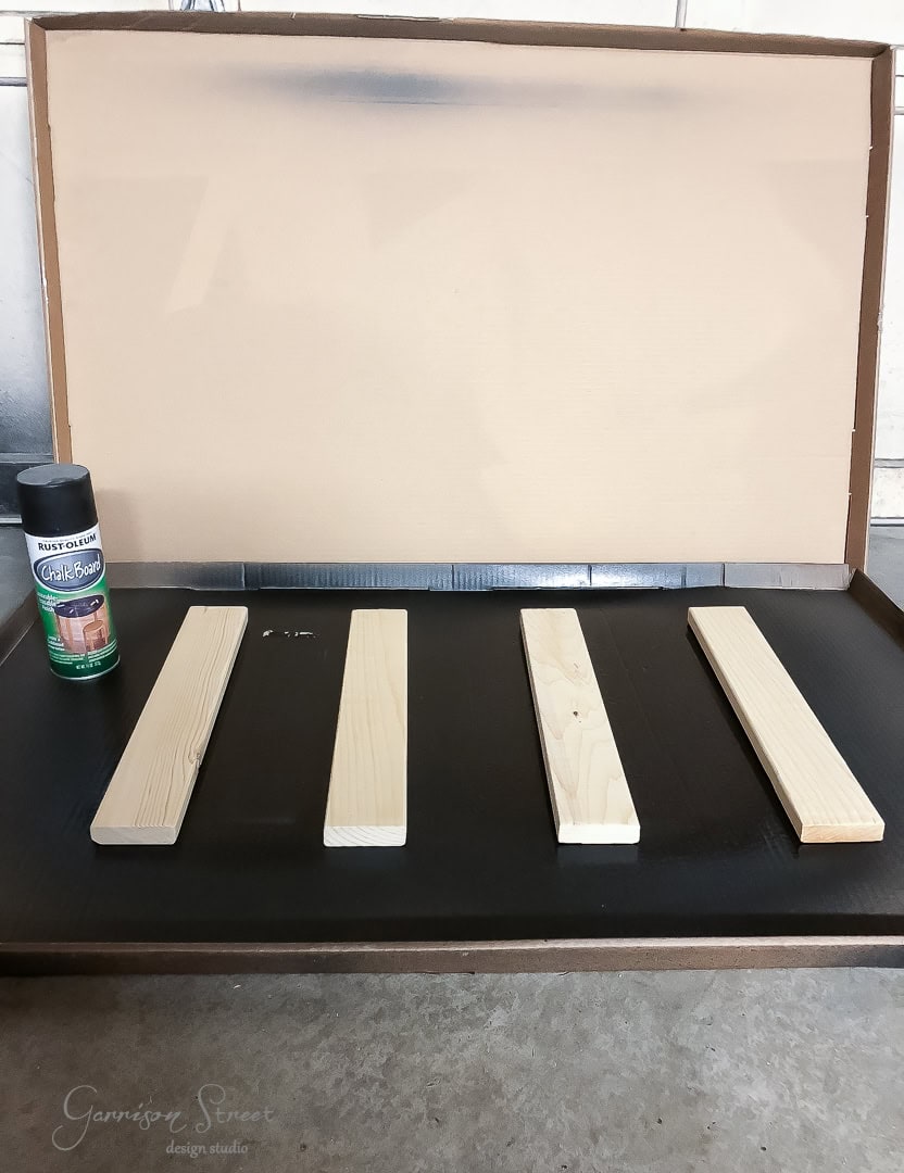 Four wooden planks are laid out on a dark tray with a can of chalkboard spray paint placed nearby. The setup appears to be for a DIY project in a workshop setting.
