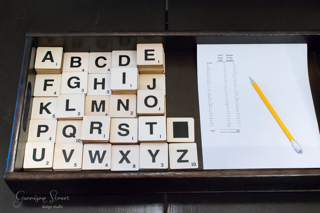 A set of wooden alphabet tiles arranged in a tray, accompanied by a blank piece of paper with tally marks and a yellow pencil placed on top.