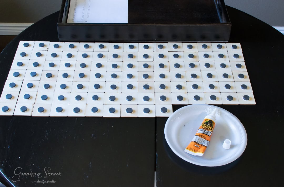 A table with a grid of small white tiles, each with a circular magnet attached. Next to the tiles, there's a white disposable plate and a tube of adhesive glue.