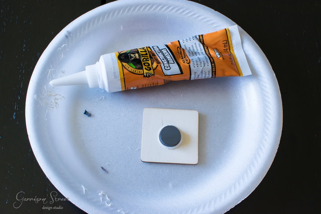 A tube of Gorilla Construction Adhesive is placed on a white paper plate beside a small wooden square with a round magnet attached. The adhesive tube is orange with black and yellow details.
