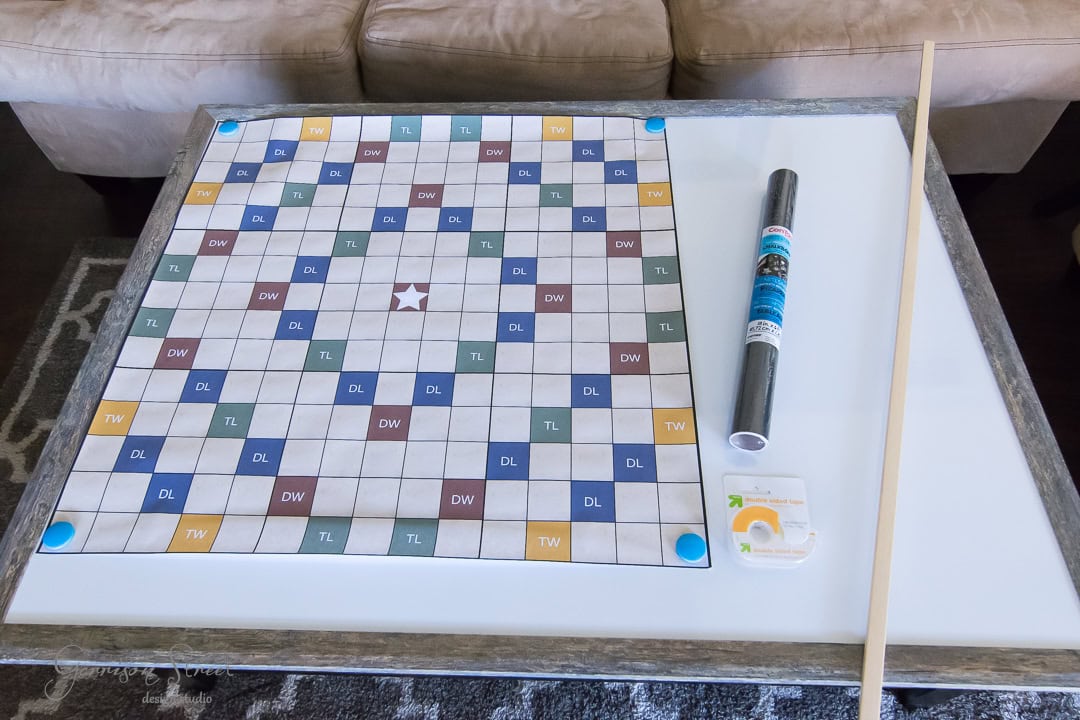 A large Scrabble board lies on a table with blue corner markers. Next to it are a black tube and a small pack with a circular object. A long wooden rod rests next to these items. A beige sofa and a patterned rug are partially visible.