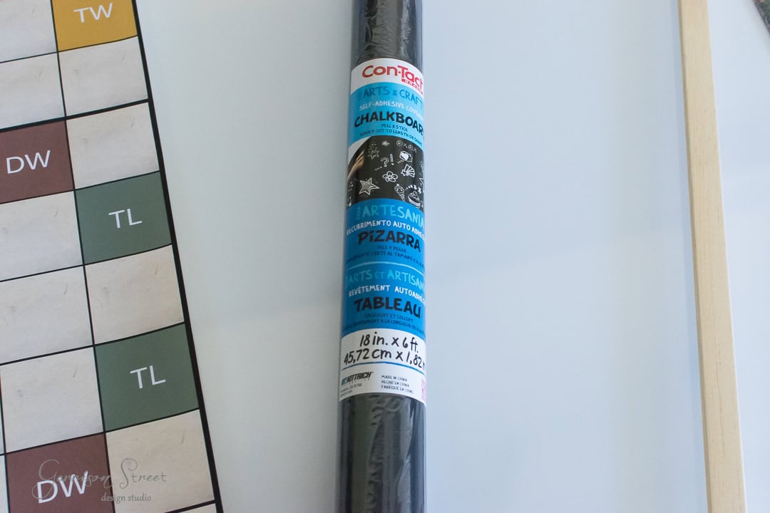 A roll of contact paper labeled as chalkboard is placed vertically. It's 18 inches wide and 6 feet long. Part of a board game or chart with "DW," "TL," and other letters is visible beside it. The background is a light surface.