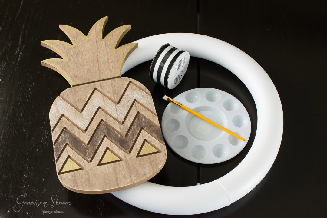 A wooden pineapple cutout with chevron patterns, a white foam wreath, a paint palette, a small paintbrush, and a spool of black and white striped ribbon are arranged on a dark surface.