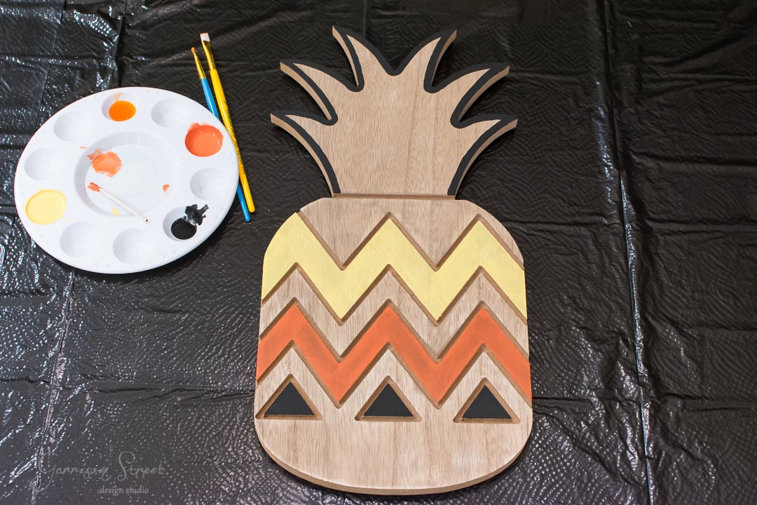 A wooden pineapple cutout with chevron patterns is placed on a black plastic-covered surface. A paint palette with orange, yellow, and black paint, along with two brushes, is beside it.