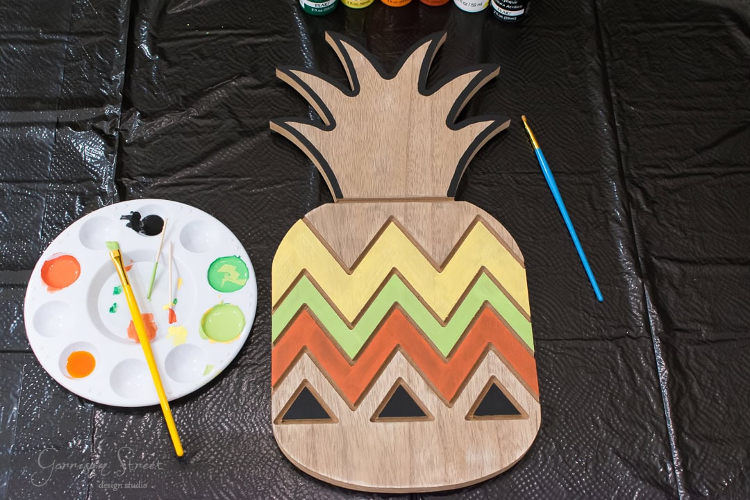 Wooden pineapple-shaped craft painted with zigzag patterns in yellow, orange, and green. A paint palette with various colors and brushes lies nearby on a black plastic surface.