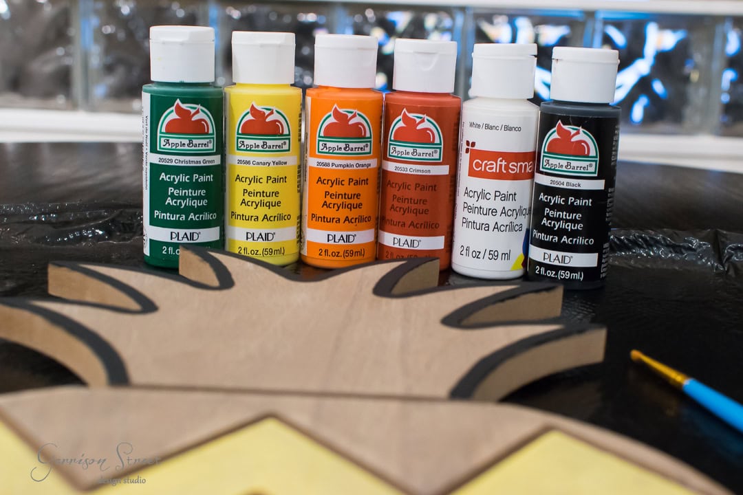 Five bottles of acrylic paint in red, orange, yellow, green, and black are lined up on a table. A craft project with a wooden cutout is partially visible in the foreground, alongside a small paintbrush.