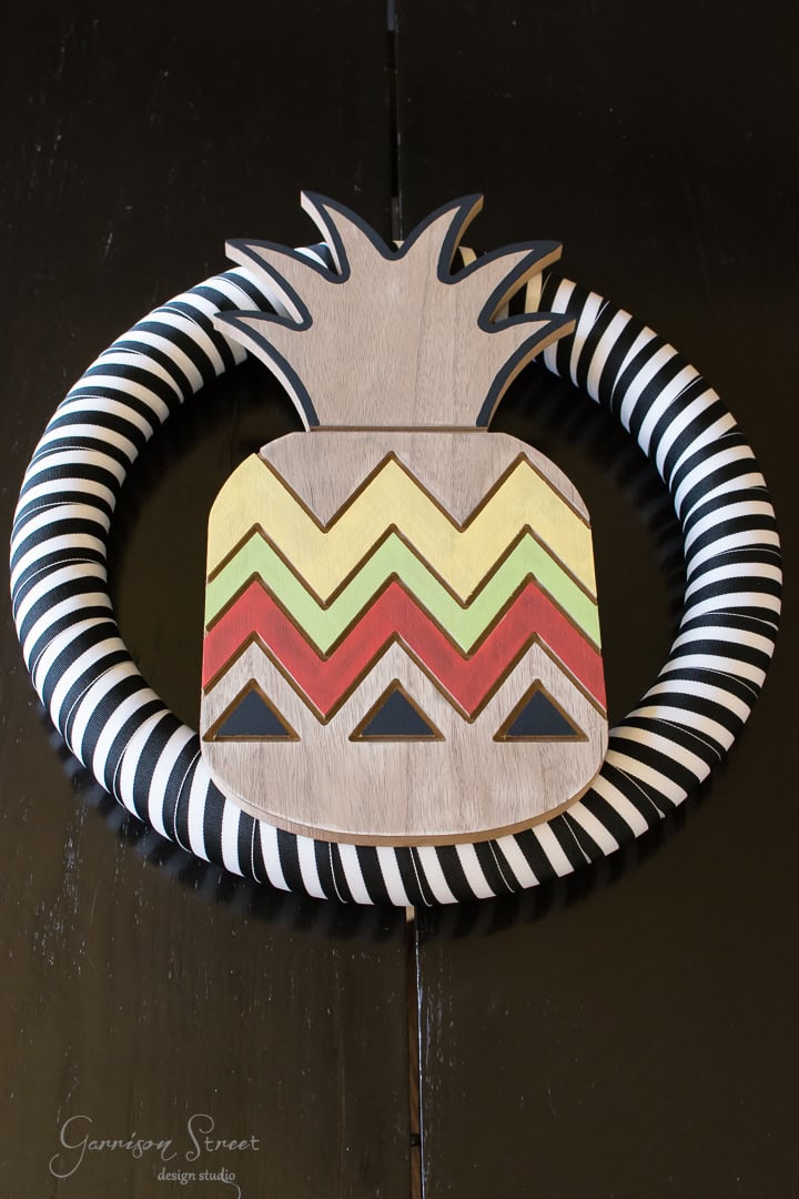 A decorative wreath featuring a wooden pineapple with zigzag and triangle patterns in yellow, red, and black, wrapped around with black and white striped cloth, displayed on a dark wooden surface.