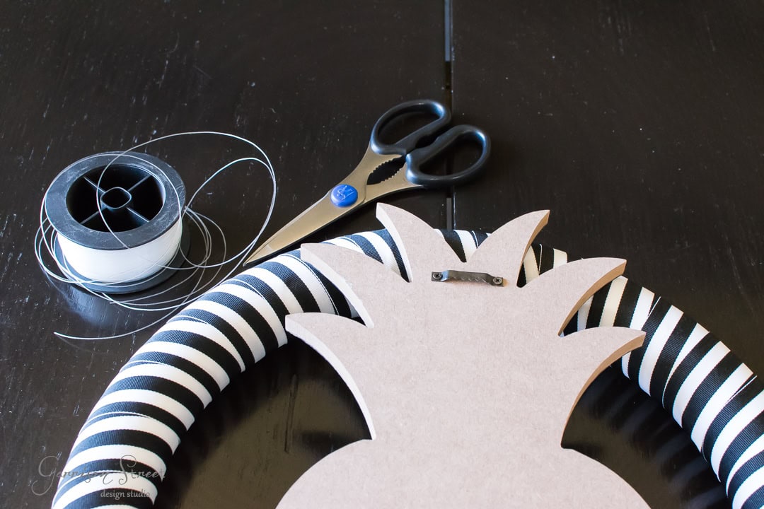 A striped black and white circle, spool of wire, scissors, and a wooden pineapple cutout with a hook are arranged on a dark surface.