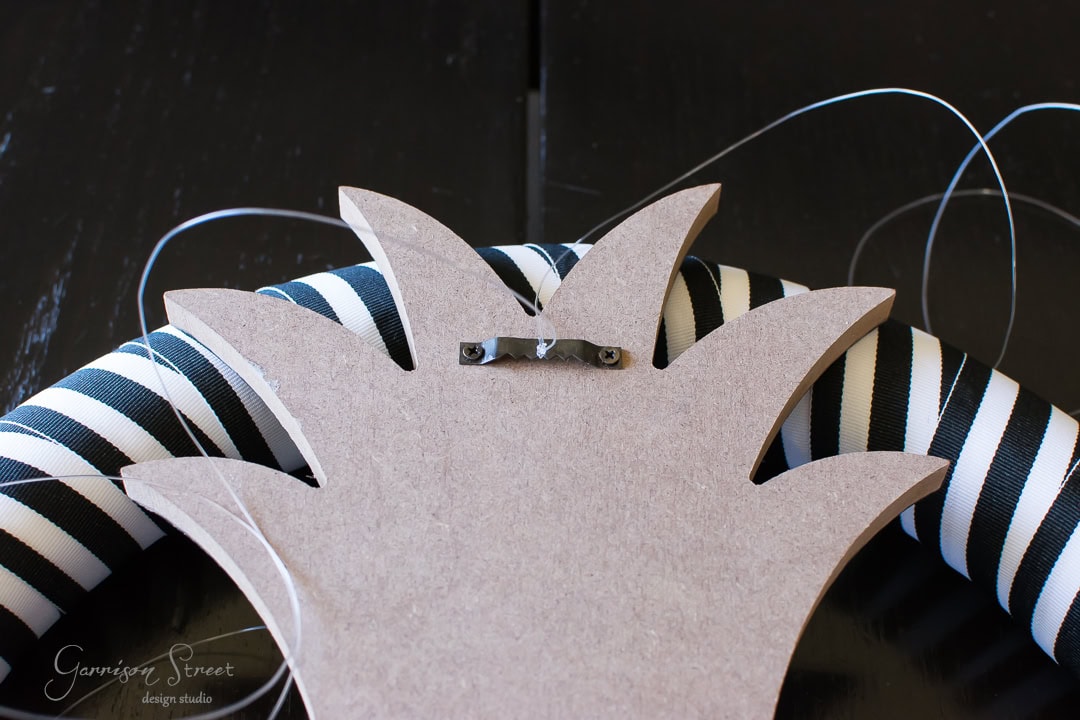 Close-up of a striped black and white wreath with a wooden back featuring a metal hook and threads. The wreath has a pineapple shape on top in unfinished wood, placed on a dark surface.