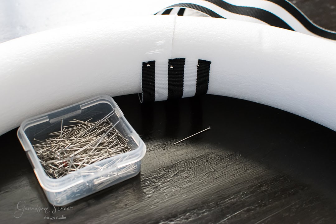 A foam ring with black and white ribbon partially wrapped around it. A container of sewing pins is nearby on a dark surface. One pin is placed separately next to the container.