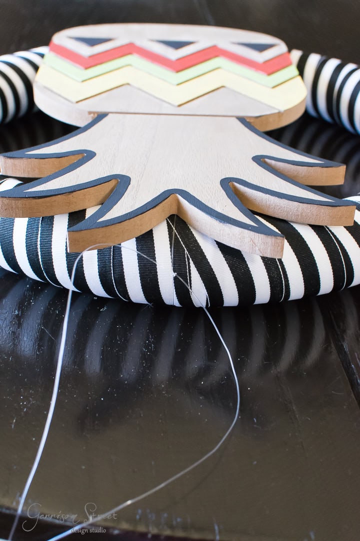 A handcrafted wooden owl with a zigzag pattern and spiral black-and-white fabric wings is shown in close-up. It lies on a dark wooden surface with visible sewing thread, indicating ongoing assembly.