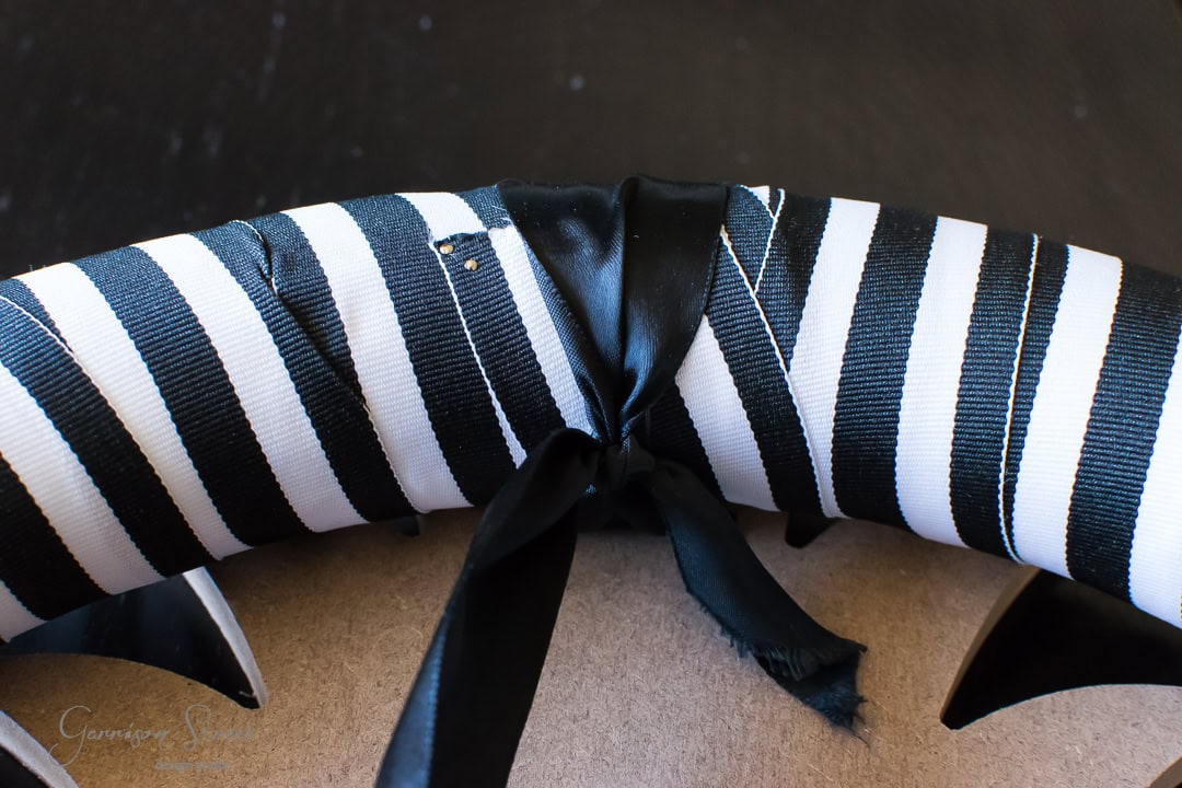 A close-up of a wreath wrapped in black and white striped fabric. A black ribbon is tied around it, forming a bow. The background is a neutral, textured surface.