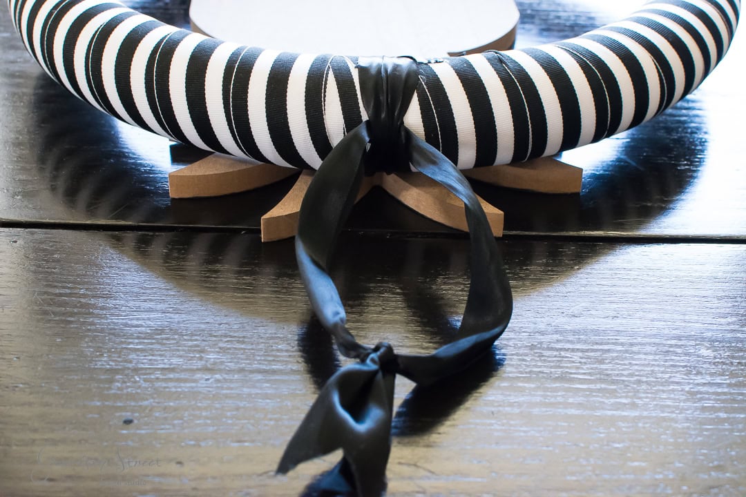 A black and white striped wreath with a black ribbon is displayed on a wooden stand. The wreath is placed on a dark, reflective surface.