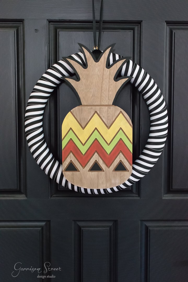 A decorative wreath on a black door features a wooden pineapple with colorful zigzag patterns in yellow, red, and green. The wreath is bordered with black and white stripes.