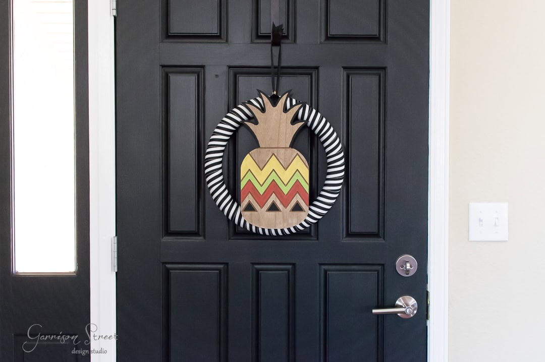 A black door features a decorative wreath with a pineapple-shaped design in yellow, red, and beige, surrounded by a black and white striped circle. The wreath hangs on the upper half of the door near the handle and lock.