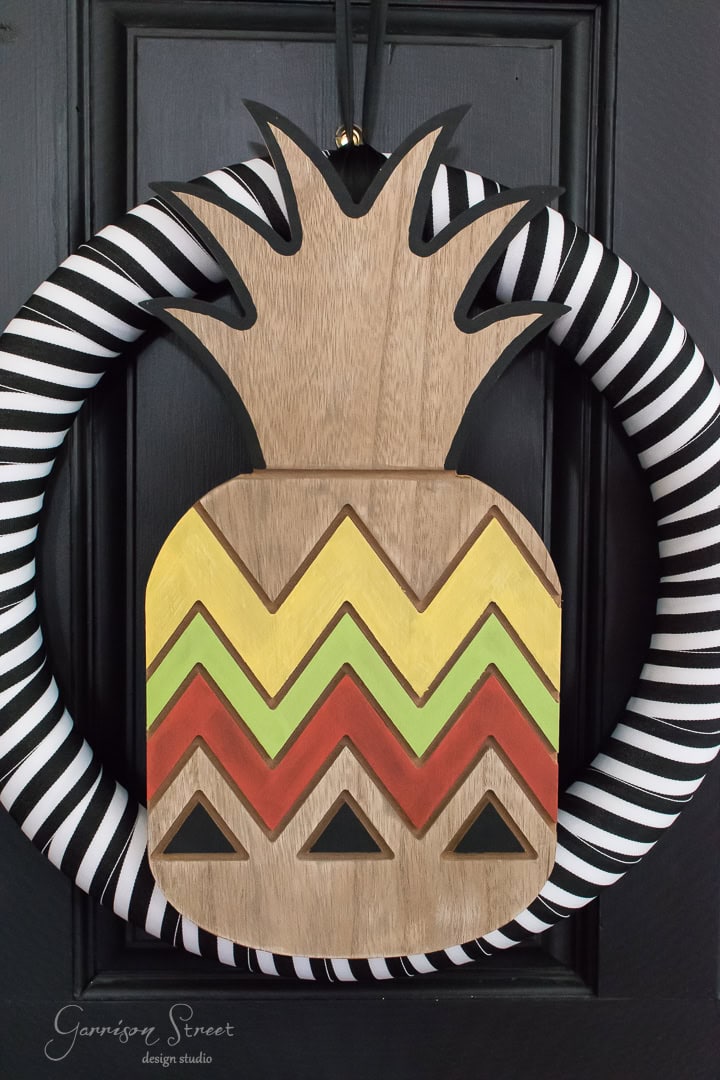 A decorative wreath on a black door features a wooden pineapple with a yellow, green, and orange chevron pattern. The wreath is wrapped in black and white stripes.