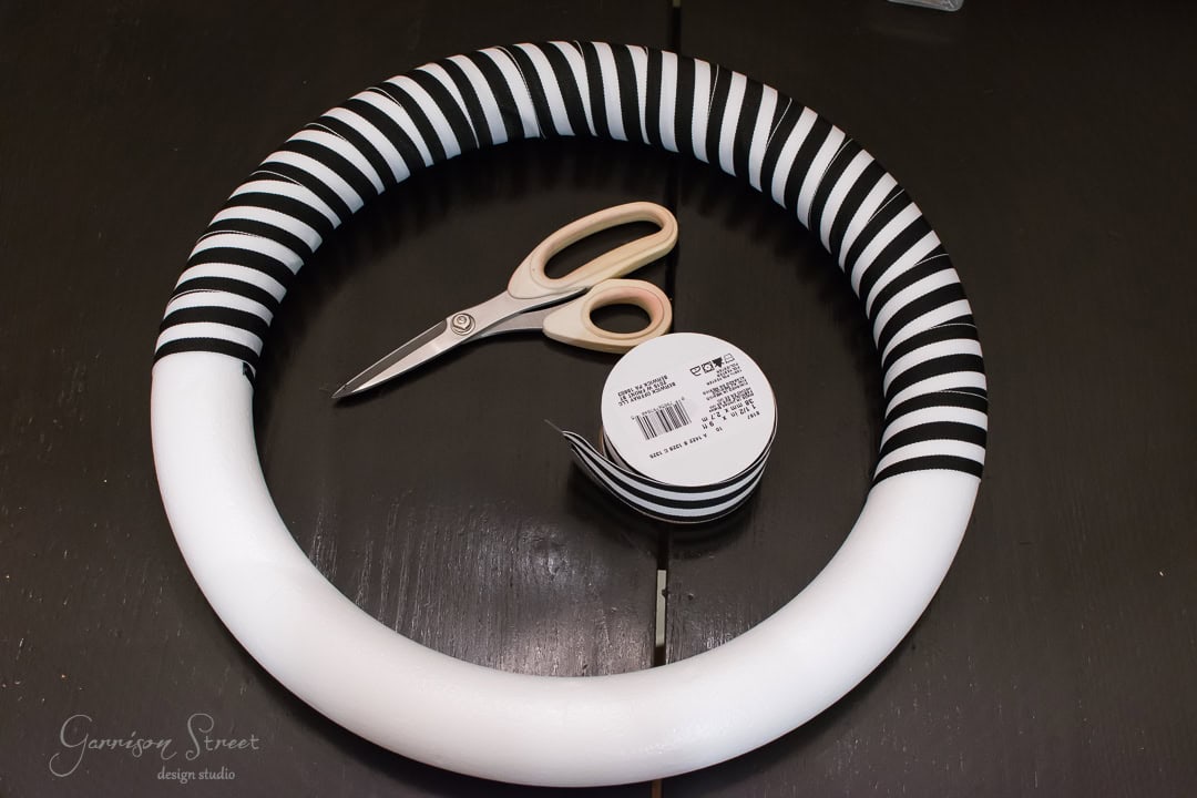 A crafting wreath covered in black and white striped fabric rests on a dark surface. Beside it are a pair of beige-handled scissors and a spool of matching black and white striped ribbon.