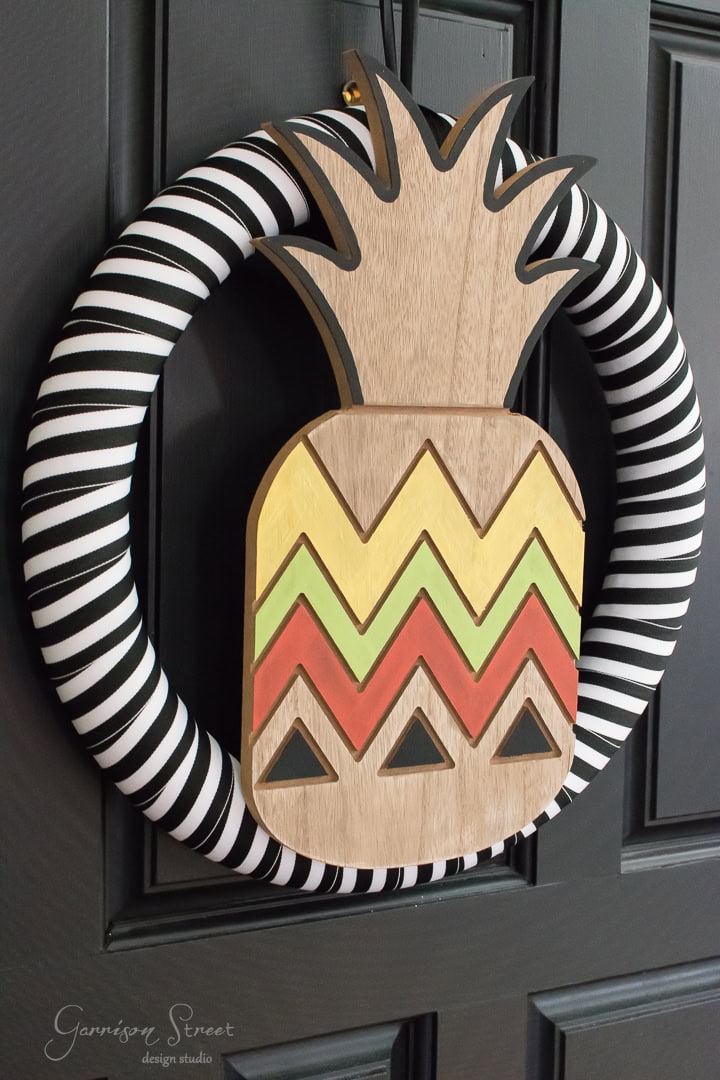 A decorative wreath hangs on a black door, featuring a wooden pineapple with colorful zigzag patterns in yellow, green, and red. The wreath is wrapped in black and white striped fabric.