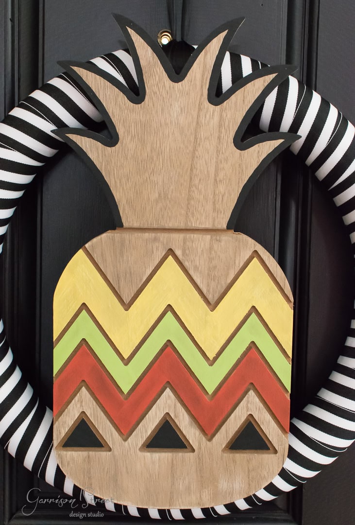 A decorative wreath featuring a wooden pineapple with chevron patterns in yellow, green, and orange. The wreath itself is wrapped in black and white striped fabric, hanging on a black door.
