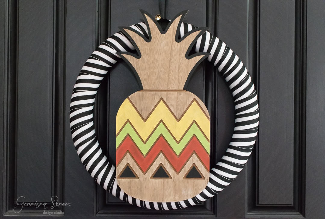 A circular wreath with a black and white striped border features a wooden pineapple centerpiece with zigzag patterns in green, yellow, and red, and small black triangles. It's hanging on a dark door.