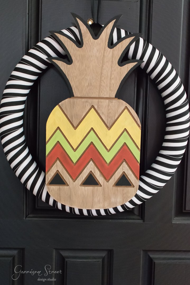 A decorative wreath features a circular black and white striped border with a wooden cutout of a pineapple in the center, displaying a zigzag pattern in yellow, orange, and green. The background is a dark painted door.