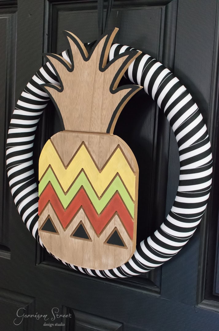 A door wreath featuring a large wooden pineapple with zigzag patterns in yellow, red, and black. The wreath is wrapped in black and white striped fabric..