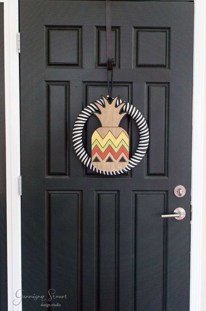 A black door with a round wreath decorated in black and white stripes, featuring a wooden pineapple with yellow, orange, and red zigzag patterns.