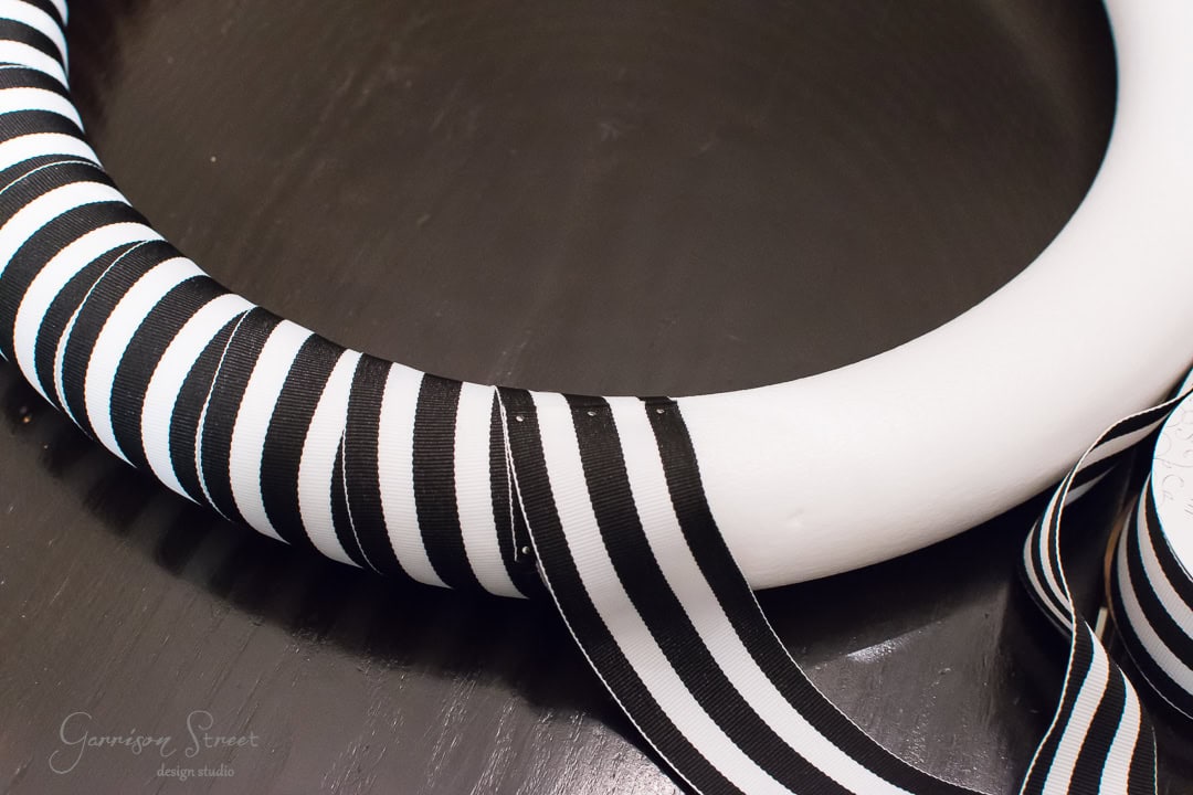 A white circular wreath form partially wrapped with a black and white striped ribbon on a dark surface. A loose end of the ribbon trails off to the right.