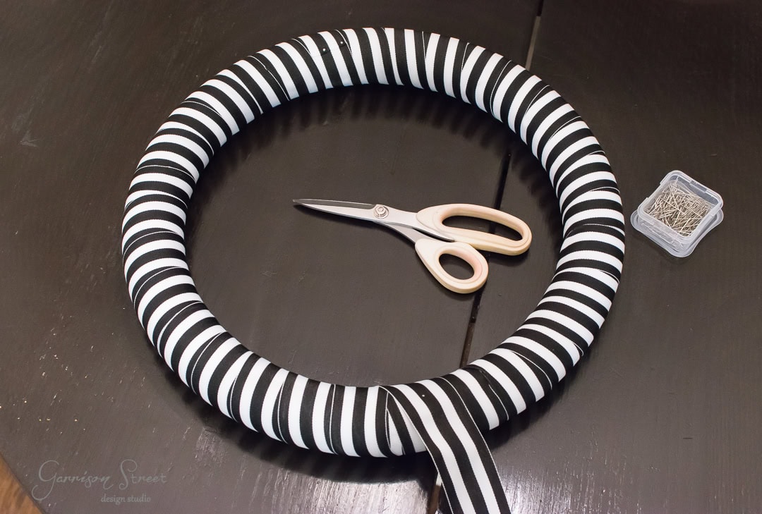 A black and white striped wreath sits on a dark table. A pair of scissors and a small container of pins are next to it. The wreath is partially wrapped with a loose ribbon.