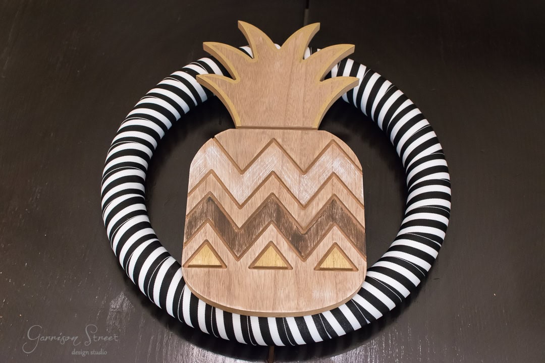 A wreath featuring a wooden pineapple shape with zigzag and triangular patterns. The wreath base is wrapped in black and white striped fabric, resting on a dark surface.