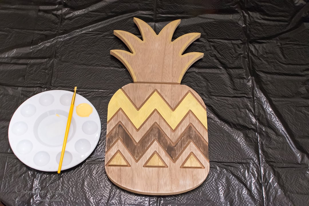 A wooden pineapple-shaped board with geometric patterns is partially painted yellow. A paintbrush with yellow paint and a palette sit nearby on a black plastic-covered surface.