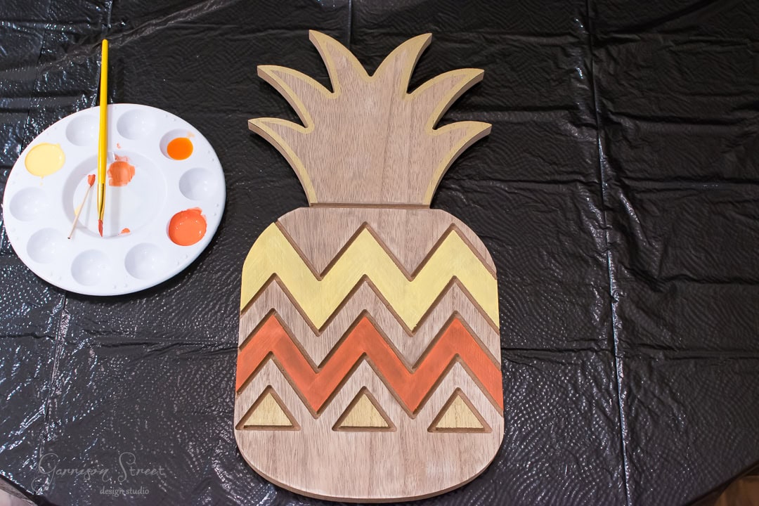 A wooden pineapple-shaped cutout lies on a table, partially painted with yellow and orange zigzag patterns. A paint palette with a brush and paint blobs is next to it. The table is covered with a black protective sheet.