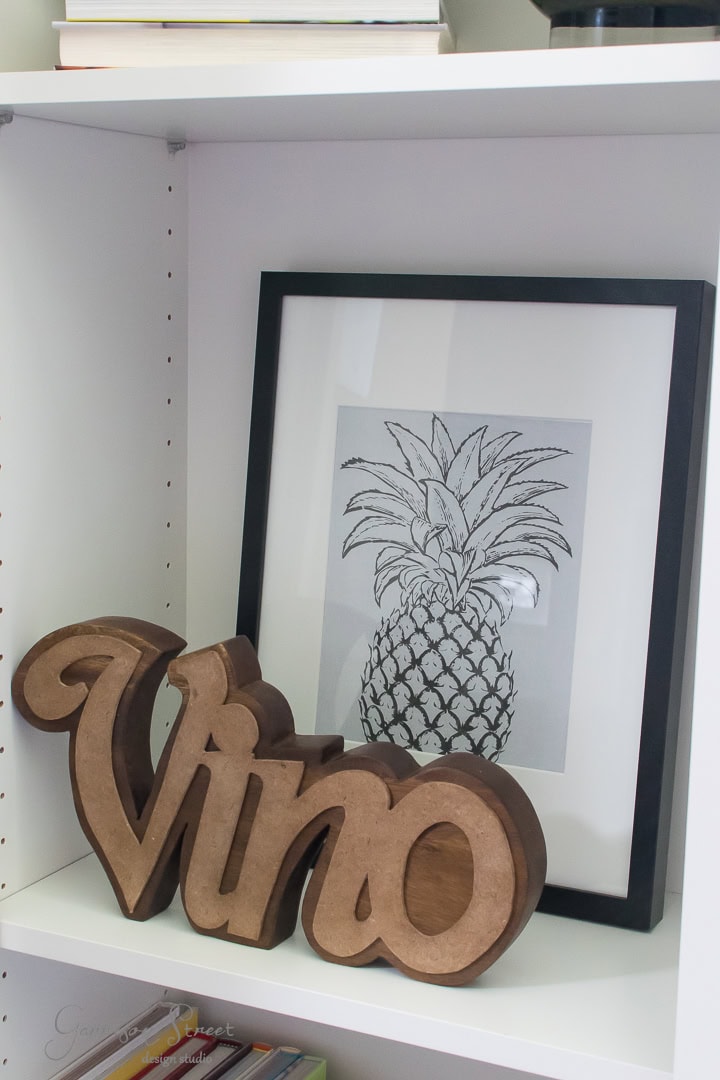 A white shelf displays a framed black and white pineapple sketch and a wooden sign saying "vino." Books are partially visible on the shelf above.