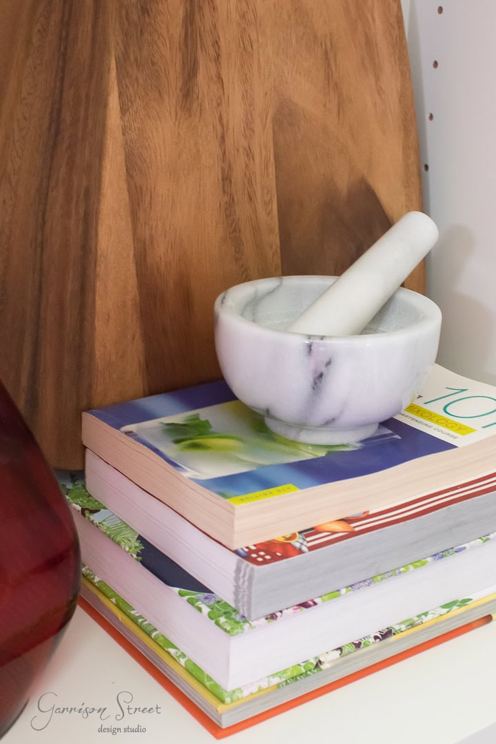 A marble mortar and pestle rests on a stack of colorful books. Behind them, a wooden cutting board is visible. The corner of a red vase peeks into the left side of the image.