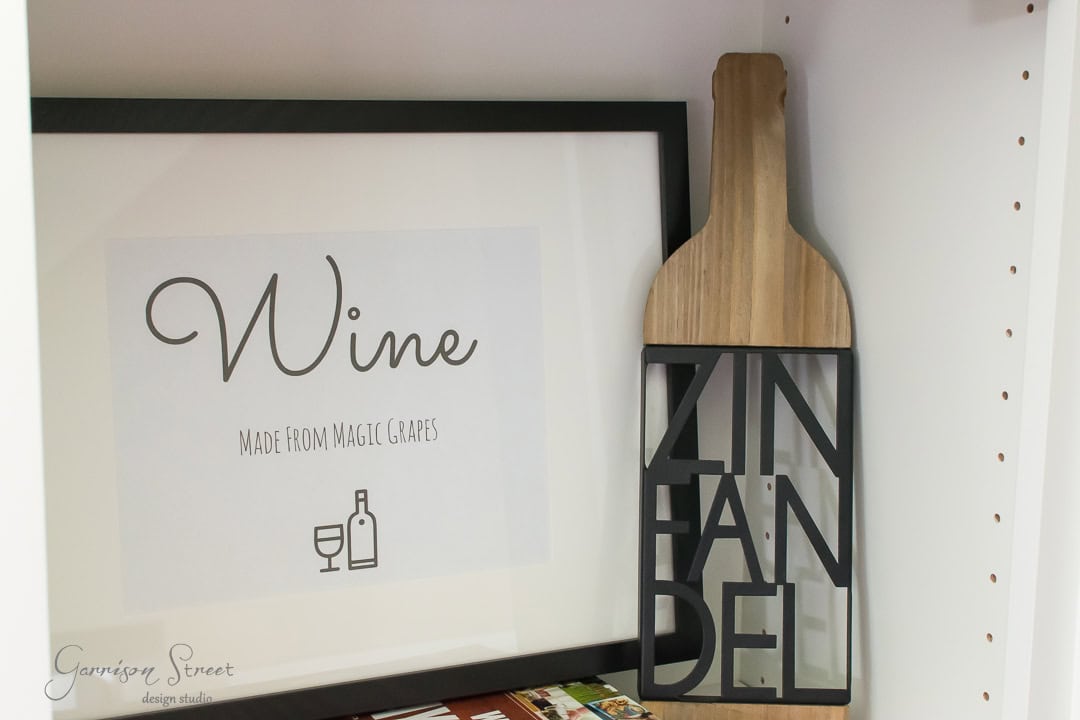 A framed artwork with the word "Wine" and "Made from Magic Grapes" displayed on a shelf. Next to it, a decorative wooden wine bottle shape with "Zinfandel" cut out. The setting is a white shelving unit.