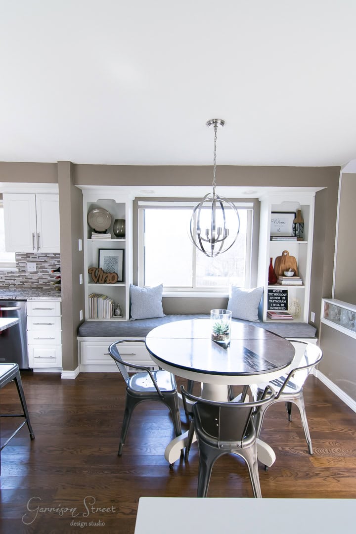 A modern dining nook with a round table surrounded by metal chairs, beneath a spherical pendant light. The nook features a window with built-in shelving on each side, decorated with books, plants, and a LOVE sign. Dark wooden floors complete the look.