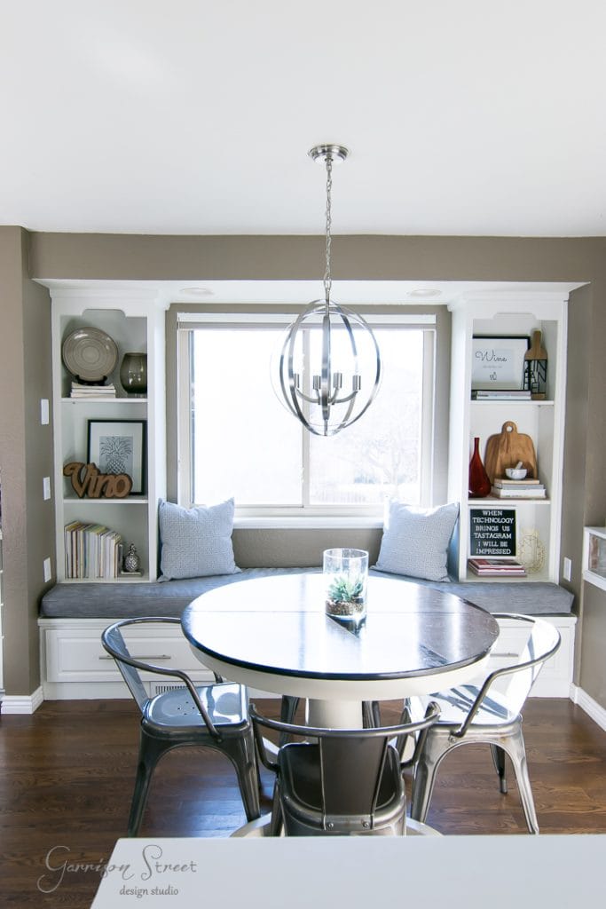 Free Printables A cozy dining nook features a round table surrounded by metal chairs, with a built-in window seat accented by gray cushions and pillows. Shelves display decorative items. A modern chandelier hangs above the table, and natural light brightens the space.