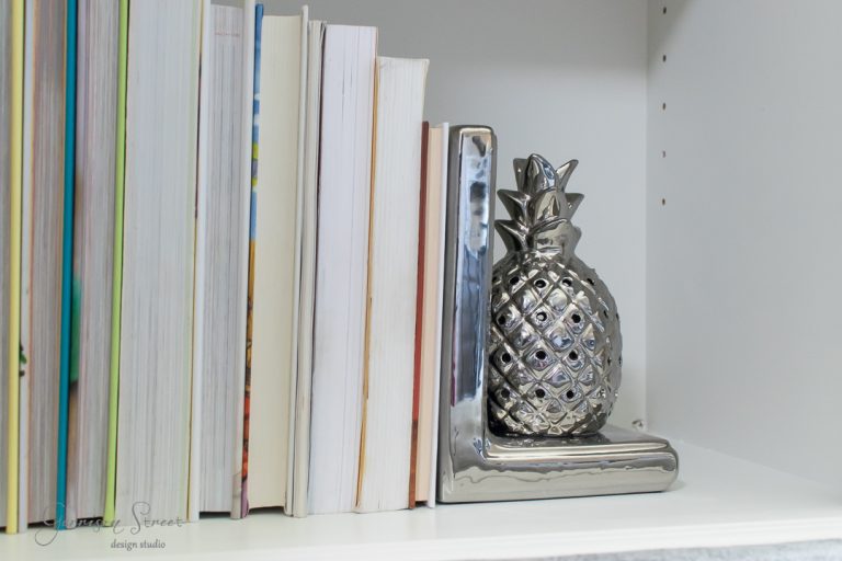 Books are lined up vertically on a shelf, held upright by a metallic pineapple-shaped bookend. The bookend has a shiny, reflective surface and is placed at the right end of the row. The shelf is white, creating a clean and modern look.