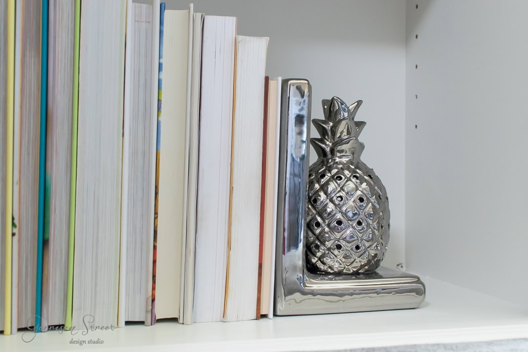 Books are lined up vertically on a shelf, held upright by a metallic pineapple-shaped bookend. The bookend has a shiny, reflective surface and is placed at the right end of the row. The shelf is white, creating a clean and modern look.
